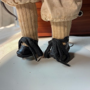 May include: A pair of black lace-up shoes with thick black laces, worn with tan ribbed socks and light tan trousers. The footwear is positioned on a circular white stand, showcasing the detailed stitching and the texture of the fabric.