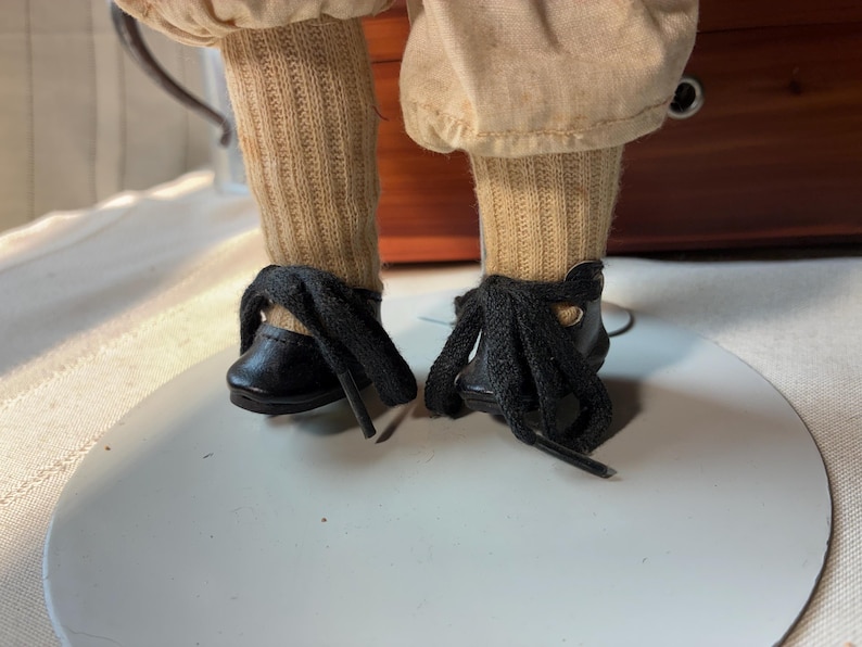 May include: A pair of black lace-up shoes with thick black laces, worn with tan ribbed socks and light tan trousers. The footwear is positioned on a circular white stand, showcasing the detailed stitching and the texture of the fabric.
