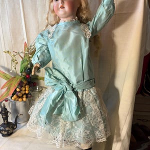 Op de afbeelding: Antieke biscuitpop met blond haar, gekleed in een lichtblauwe satijnen jurk met een grote strik en een witte kanten rok. De pop heeft een geschilderd gezicht en poseert met &eacute;&eacute;n arm omhoog. Zwarte schoenen maken de outfit af.
