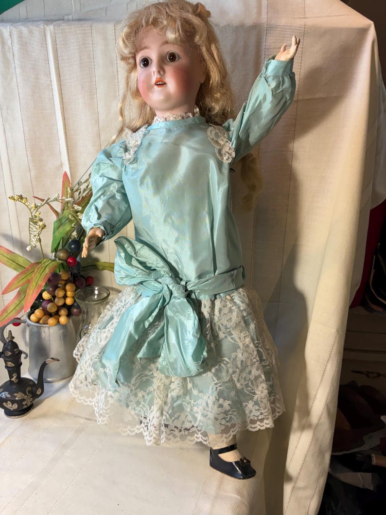 Op de afbeelding: Antieke biscuitpop met blond haar, gekleed in een lichtblauwe satijnen jurk met een grote strik en een witte kanten rok. De pop heeft een geschilderd gezicht en poseert met &eacute;&eacute;n arm omhoog. Zwarte schoenen maken de outfit af.