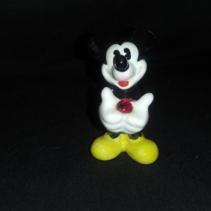 May include: A small, ceramic Mickey Mouse figurine. The figure has black ears, a black nose, and a red mouth. It wears white gloves and yellow shoes. A red gem is held in its hands.