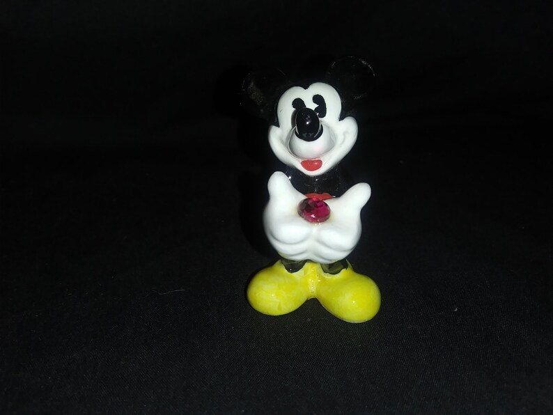 May include: A small, ceramic Mickey Mouse figurine. The figure has black ears, a black nose, and a red mouth. It wears white gloves and yellow shoes. A red gem is held in its hands.