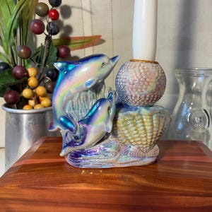 May include: Iridescent ceramic dolphin figurine with two dolphins leaping from the water. The figurine is set on a seashell base and has a candle holder. The dolphins have blue and purple hues.