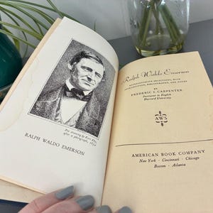 May include: An open book featuring a portrait of Ralph Waldo Emerson and the title "Ralph Waldo Emerson." The book's pages are aged, with text from the American Book Company visible. The cover is a light tan color.