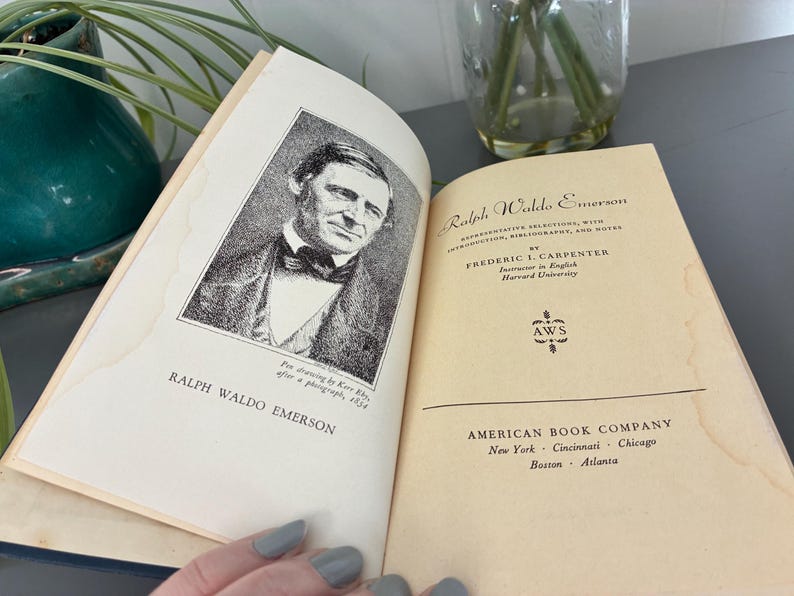 May include: An open book featuring a portrait of Ralph Waldo Emerson and the title "Ralph Waldo Emerson." The book's pages are aged, with text from the American Book Company visible. The cover is a light tan color.