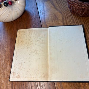 May include: An open book with aged, off-white pages and a black cover. The pages show signs of age with brown spotting. The book is lying on a wooden surface, with a decorative seashell and a woven basket in the background.