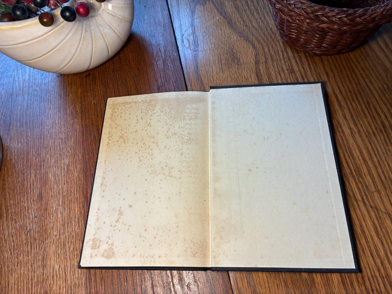 May include: An open book with aged, off-white pages and a black cover. The pages show signs of age with brown spotting. The book is lying on a wooden surface, with a decorative seashell and a woven basket in the background.
