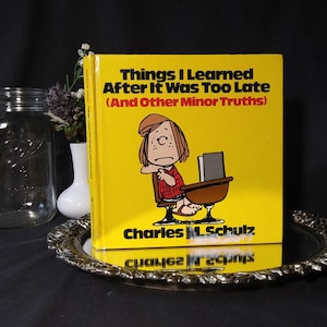 May include: A yellow book titled "Things I Learned After It Was Too Late (And Other Minor Truths)" by Charles M. Schulz. The cover features a cartoon character sitting at a desk. The book is on a decorative silver tray.