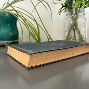 May include: A vintage book with a dark teal cover and gold accents. The pages are aged with a golden hue. The book is resting on a reflective surface, with a potted plant and a jar of flowers in the background.