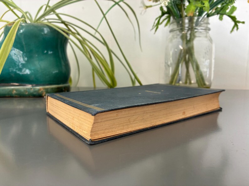 May include: A vintage book with a dark teal cover and gold accents. The pages are aged with a golden hue. The book is resting on a reflective surface, with a potted plant and a jar of flowers in the background.