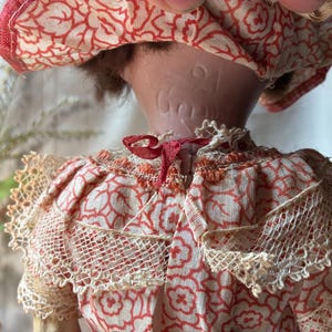 May include: A doll wearing a cream-colored fabric garment with a red floral pattern and lace trim. The back of the doll's neck features an embossed inscription that reads 21 and Gema. The doll has brown hair and a matching red and cream hat.