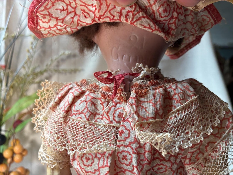 May include: A doll wearing a cream-colored fabric garment with a red floral pattern and lace trim. The back of the doll's neck features an embossed inscription that reads 21 and Gema. The doll has brown hair and a matching red and cream hat.