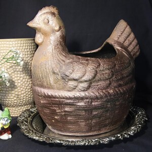 May include: A large, bronze-colored chicken-shaped planter with a textured surface. The planter sits on a silver-colored, ornate tray. A white vase with flowers and a small figurine are also in the image.