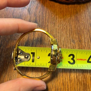 May include: A gold-colored bangle bracelet with a decorative center, measured at approximately 2.5 inches in diameter. The bracelet is being measured with a yellow tape measure, showing the size of the jewelry.