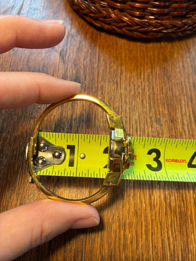 May include: A gold-colored bangle bracelet with a decorative center, measured at approximately 2.5 inches in diameter. The bracelet is being measured with a yellow tape measure, showing the size of the jewelry.