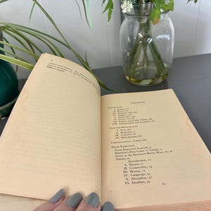 May include: An open book with aged, yellowed pages, displaying the table of contents and text. The book is on a gray surface, with a green plant and a glass jar with flowers in the background. The text includes "Contents" and "Nature, 10".