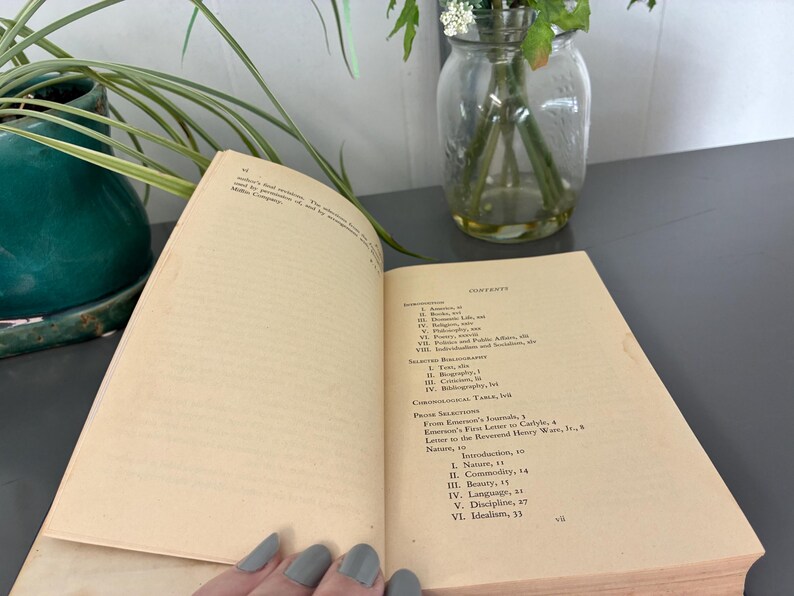 May include: An open book with aged, yellowed pages, displaying the table of contents and text. The book is on a gray surface, with a green plant and a glass jar with flowers in the background. The text includes "Contents" and "Nature, 10".