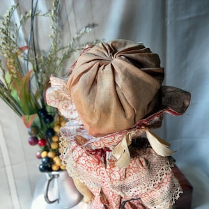 May include: A doll wearing a tan fabric bonnet with a gathered crown and a ruffled brim featuring a red and white floral pattern. The doll is dressed in a matching red and white floral print garment with lace trim and a tied ribbon under the chin.