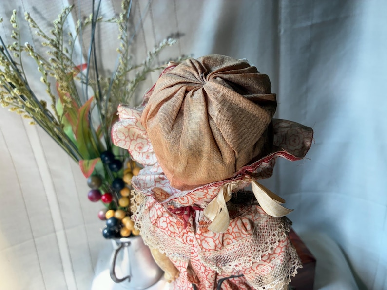 May include: A doll wearing a tan fabric bonnet with a gathered crown and a ruffled brim featuring a red and white floral pattern. The doll is dressed in a matching red and white floral print garment with lace trim and a tied ribbon under the chin.