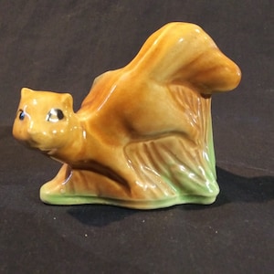 May include: A ceramic squirrel figurine in shades of brown, tan, and green. The squirrel is posed in a sitting position, with its tail curved upwards. The figurine has black eyes and is set on a green base.