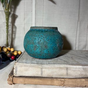 May include: A round, turquoise ceramic vase with a textured surface and a dark rim. The vase is displayed on top of two antique books. The vase is approximately 6 inches in diameter.