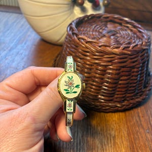 May include: A vintage gold-toned watch with a white face and floral design. The watch band is white with floral accents. The watch is held in front of a woven basket and a decorative ceramic bowl.