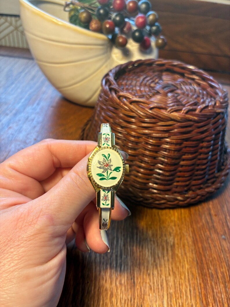 May include: A vintage gold-toned watch with a white face and floral design. The watch band is white with floral accents. The watch is held in front of a woven basket and a decorative ceramic bowl.