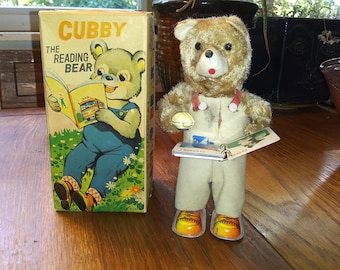 Può includere: Giocattolo vintage Cubby l'orso lettore con scatola. La scatola presenta un orso dei cartoni animati che legge un libro. L'orso giocattolo indossa un completo bianco, uno stetoscopio rosso e tiene in mano un libro. L'orso ha scarpe gialle e arancioni.