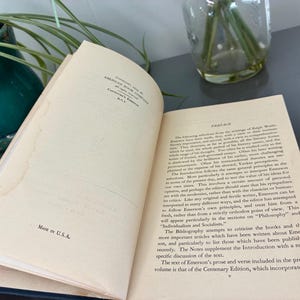 May include: An open book with text on aged pages. The text includes the word "Preface" and "MADE IN U.S.A." The book is part of the Centenary Edition. The book is on a gray surface with a green plant and a glass vase in the background.