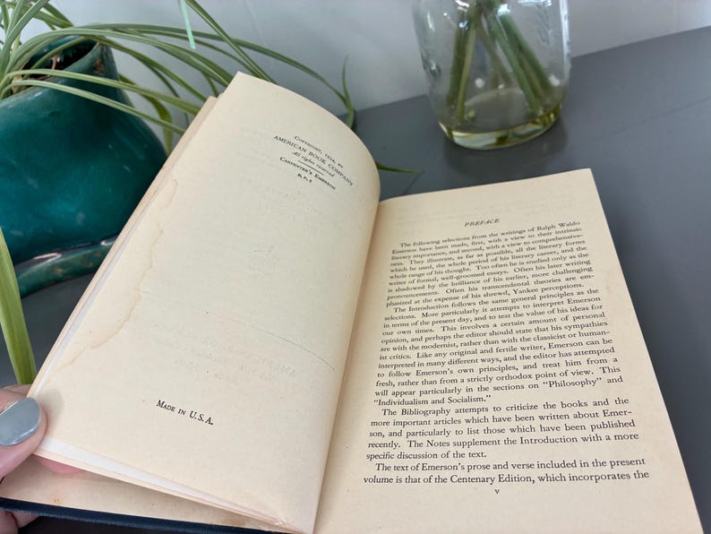 May include: An open book with text on aged pages. The text includes the word "Preface" and "MADE IN U.S.A." The book is part of the Centenary Edition. The book is on a gray surface with a green plant and a glass vase in the background.