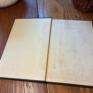 May include: An open book with aged, off-white pages and a black spine. The pages show signs of age with brown spotting. The book is lying on a wooden surface, with a white object and a wicker basket in the background.