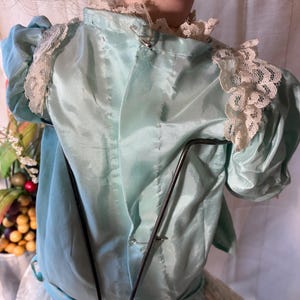 Op de afbeelding: Een vintage poppenjurk in lichtblauwe satijnen stof met delicate kanten afwerking op de schouders en halslijn. De jurk heeft korte, gepofte mouwen en is vastgemaakt met veiligheidsspelden. De rok van de pop is zichtbaar, gemaakt van witte kant.