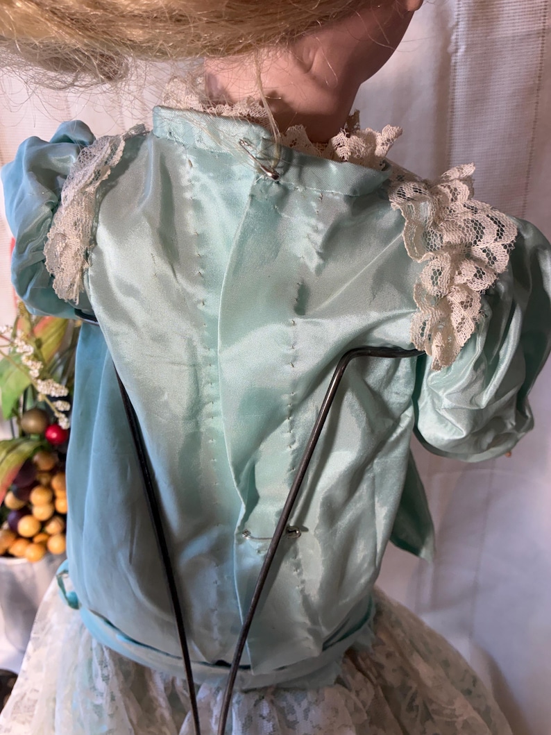 Op de afbeelding: Een vintage poppenjurk in lichtblauwe satijnen stof met delicate kanten afwerking op de schouders en halslijn. De jurk heeft korte, gepofte mouwen en is vastgemaakt met veiligheidsspelden. De rok van de pop is zichtbaar, gemaakt van witte kant.