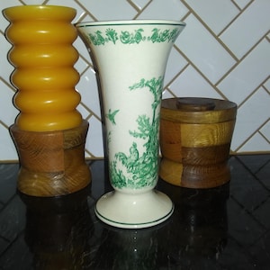 Puede incluir: Tres portavelas de madera, uno con una vela amarilla, otro con un jarrón blanco con diseños florales verdes y otro con tapa. Los portavelas están sobre una superficie negra.