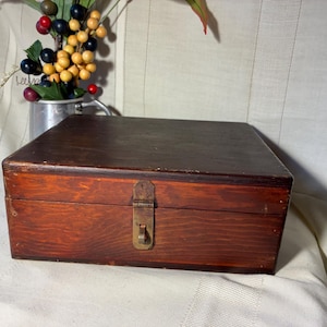 May include: A rectangular wooden storage box with a dark brown stained finish and visible wood grain. The box features a hinged lid and a central brass-toned metal latch mechanism on the front panel for securing the closure of the container.