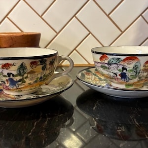 May include: Two vintage teacups and saucers. The cups and saucers are white with a black trim and feature a colorful landscape scene with figures. The cups have handles and sit on matching saucers.