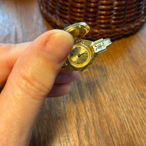 May include: A gold-toned ring watch with a hinged cover, featuring a round gold face with black numerals and hands. The watch has a floral design on the band. The watch is held in a hand.