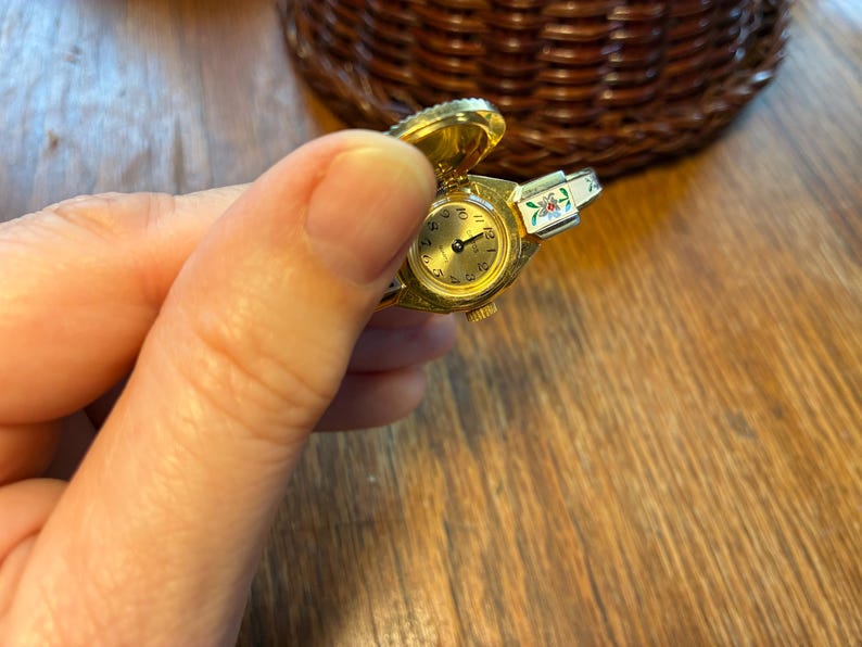 May include: A gold-toned ring watch with a hinged cover, featuring a round gold face with black numerals and hands. The watch has a floral design on the band. The watch is held in a hand.