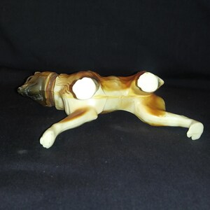 May include: A brown and white plastic toy dog figurine. The dog is lying on its belly with its legs outstretched. The dog has a brown head and tail, and white body and legs.