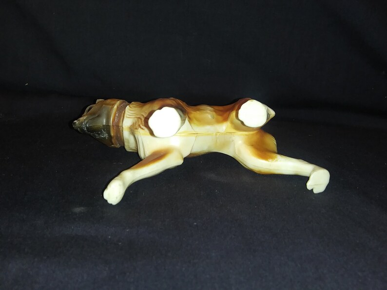 May include: A brown and white plastic toy dog figurine. The dog is lying on its belly with its legs outstretched. The dog has a brown head and tail, and white body and legs.