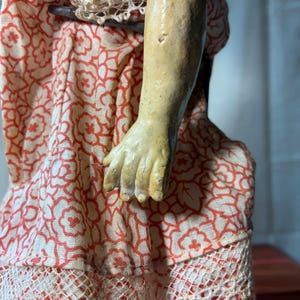 May include: A doll arm and hand with a textured, light tan finish, positioned against a dress made of cream-colored fabric featuring a repeating orange floral pattern and a white lace trim along the bottom edge of the garment.
