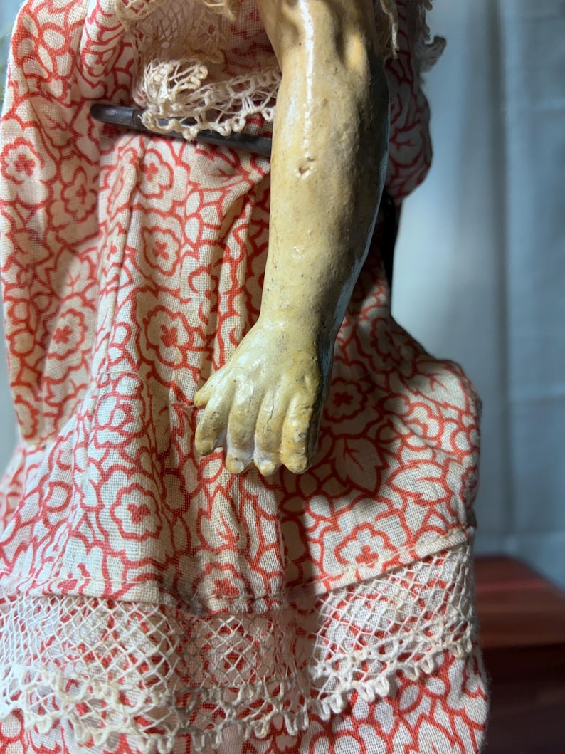 May include: A doll arm and hand with a textured, light tan finish, positioned against a dress made of cream-colored fabric featuring a repeating orange floral pattern and a white lace trim along the bottom edge of the garment.