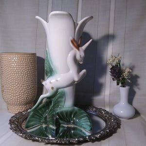 May include: A white ceramic vase with a deer figurine, green leaves, and a silver tray. A beige textured vase and a small white vase with dried flowers are also present. The deer has brown spots and ears.