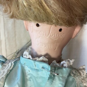 Op de afbeelding: Een vintage porseleinen pop met blond haar en een lichtblauwe jurk met witte kanten afwerking. De nek van de pop is gemarkeerd met de tekst "Queen Louise Germany". Het hoofd van de pop is iets naar voren gekanteld.