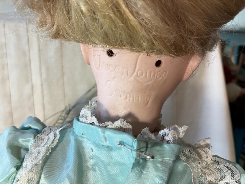 Op de afbeelding: Een vintage porseleinen pop met blond haar en een lichtblauwe jurk met witte kanten afwerking. De nek van de pop is gemarkeerd met de tekst "Queen Louise Germany". Het hoofd van de pop is iets naar voren gekanteld.