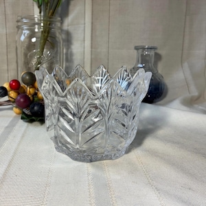 May include: A clear, faceted glass bowl with a leaf design. The bowl has a scalloped rim and is sitting on a white surface. Other objects in the background include a glass jar, a small blue vase, and decorative berries.