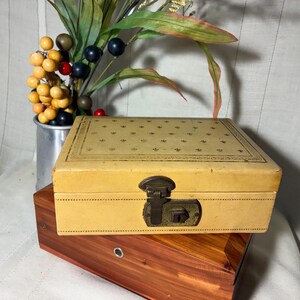May include: A rectangular cream-colored box with a repeating fleur-de-lis pattern on the lid and a dark metal latch closure. The box rests on top of a rectangular wooden box with a visible metal keyhole and a distinct reddish-brown wood grain.