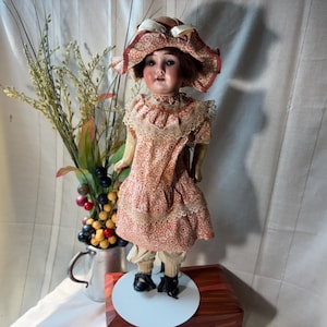 May include: A vintage doll with a painted face, brown hair, and a wide-brimmed hat. The doll wears a peach-colored dress with a repeating circular pattern, lace trim, and light-colored trousers. It stands on a white circular base with black shoes.
