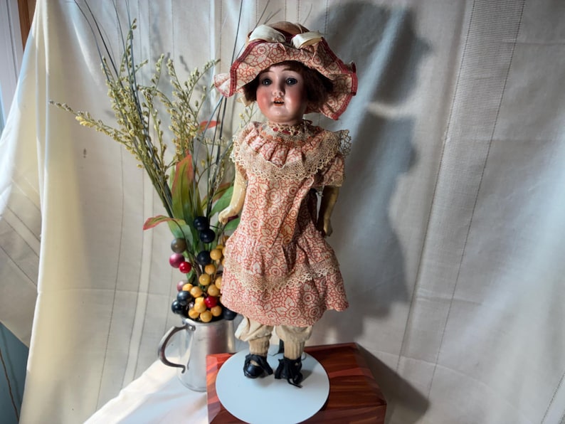May include: A vintage doll with a painted face, brown hair, and a wide-brimmed hat. The doll wears a peach-colored dress with a repeating circular pattern, lace trim, and light-colored trousers. It stands on a white circular base with black shoes.