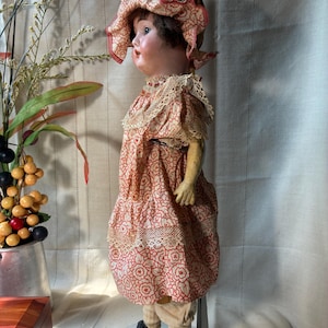 May include: A vintage doll with a painted face and dark hair, wearing a ruffled bonnet and a dress featuring a red and white floral pattern with lace trim. The doll has light-colored leggings and black shoes, standing on a metal display stand.